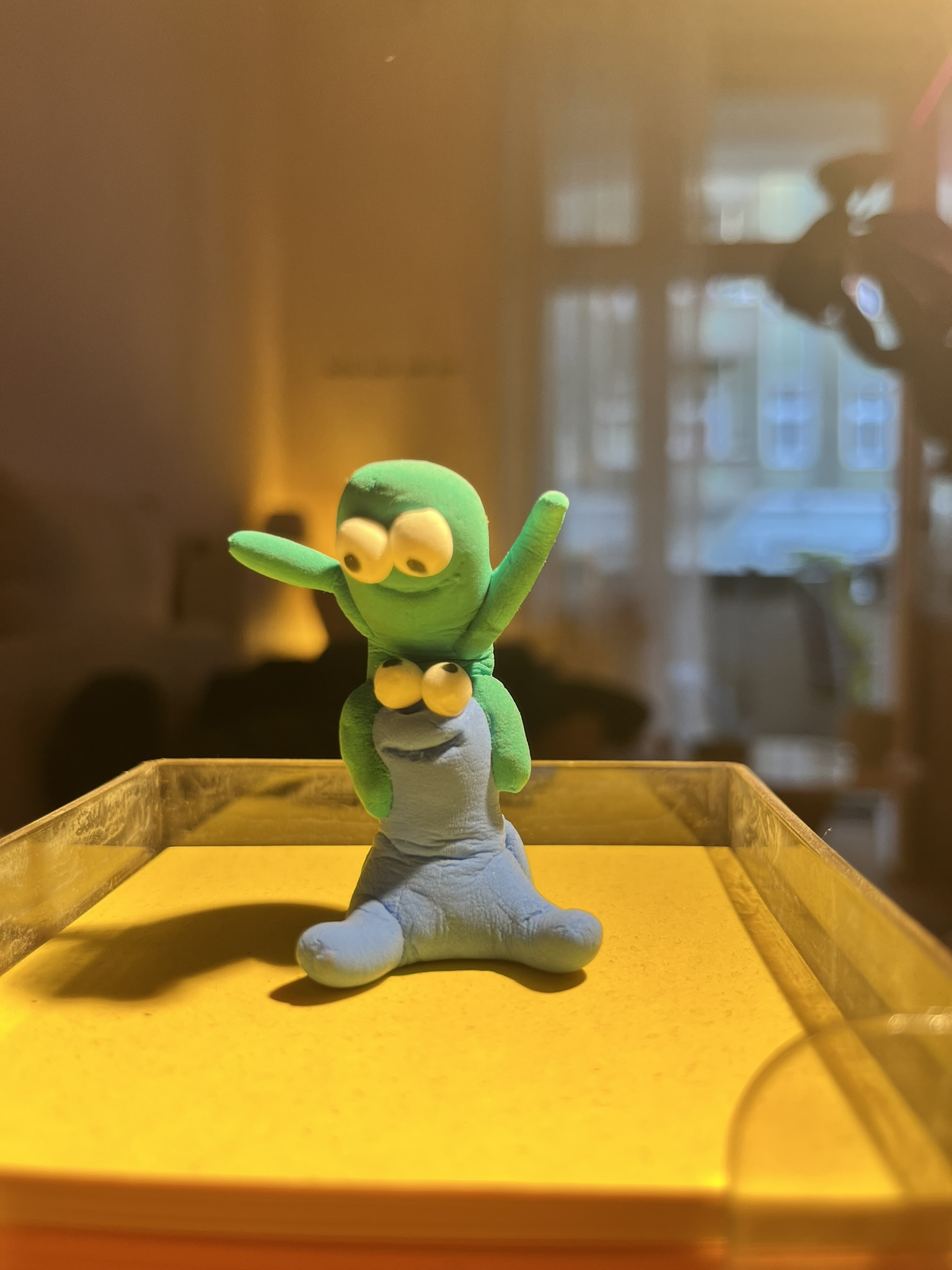 Grüner Squig sitzt huckepack auf blauem Squig auf goldenem Tablett am Fenster